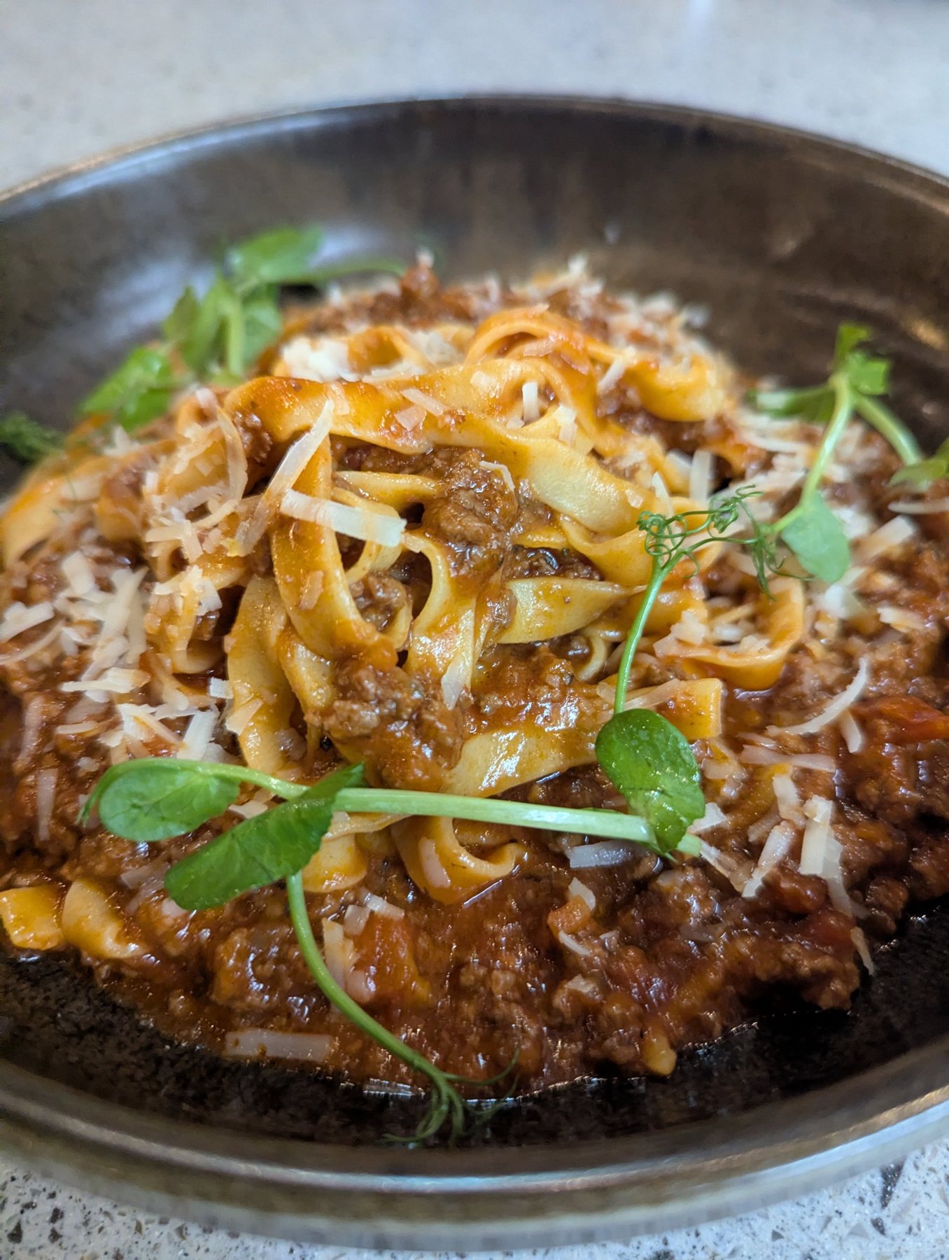 Pappardelle tossed through a dark bolognese ragù, with grated cheese and pea shoots